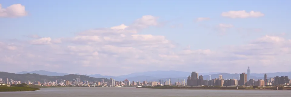 淡藍天空與粉色雲層下，河岸另一側的台北城市天際線。